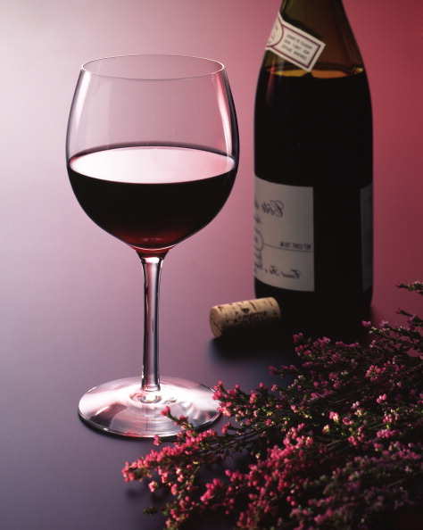 Still life of a glass of wine standing next to a wine bottle, a cork and several stalks of pink flowers before a seamless multicolored background