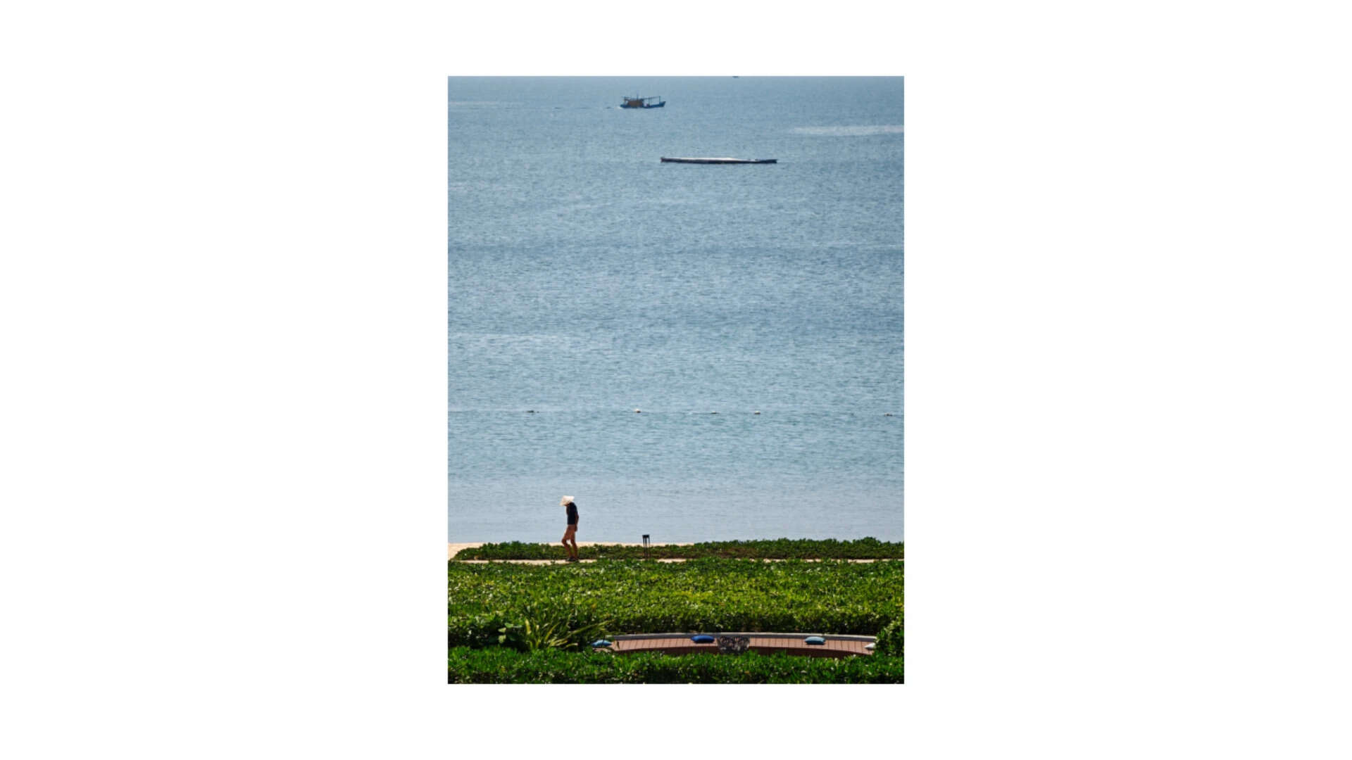 The telephoto's compression at work—a person on the beachfront, the sea, and the boats beyond, all flattened into a single layered frame