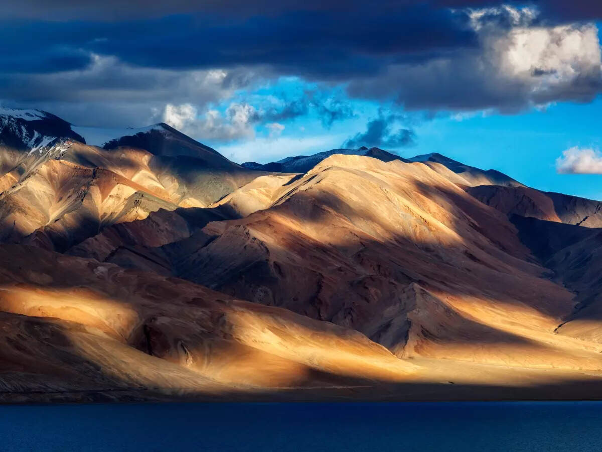 Golden sunlit mountains with dramatic cloud shadows reflecting over a blue high-altitude lake in Ladakh